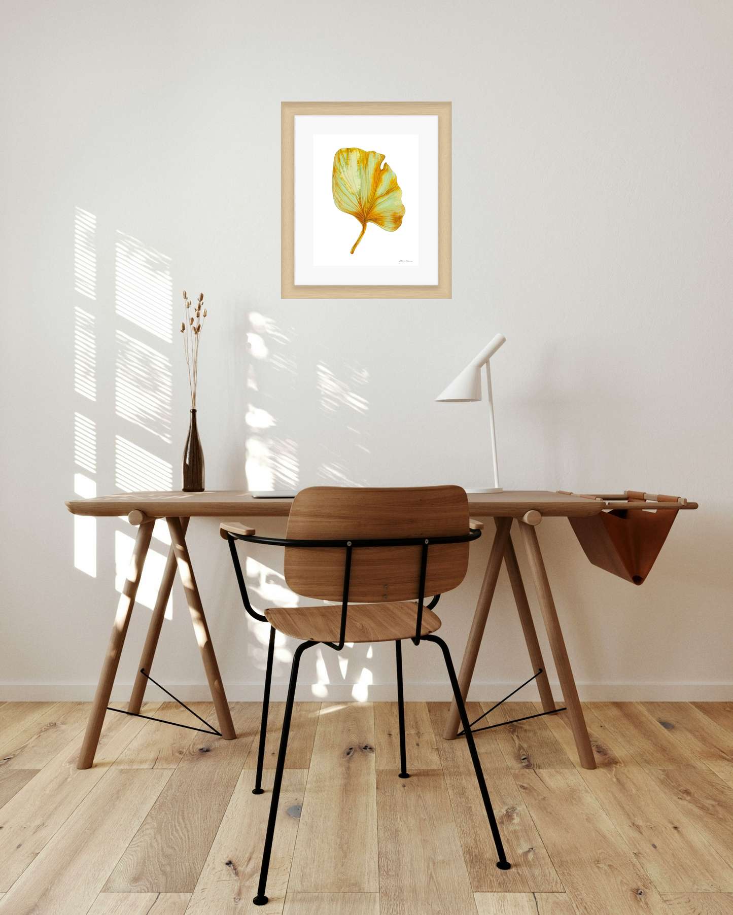 Wooden desk with chair in a room with a framed artwork of a yellow and green leaf on a white background on the wall.