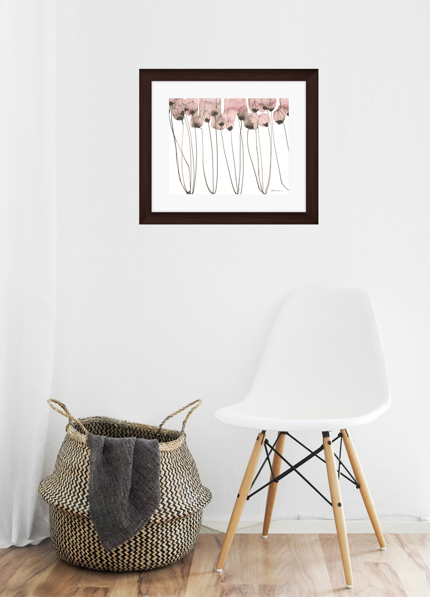 Framed artwork of pink flowers with long stems on a white background on a wall with a chair and woven basket below.