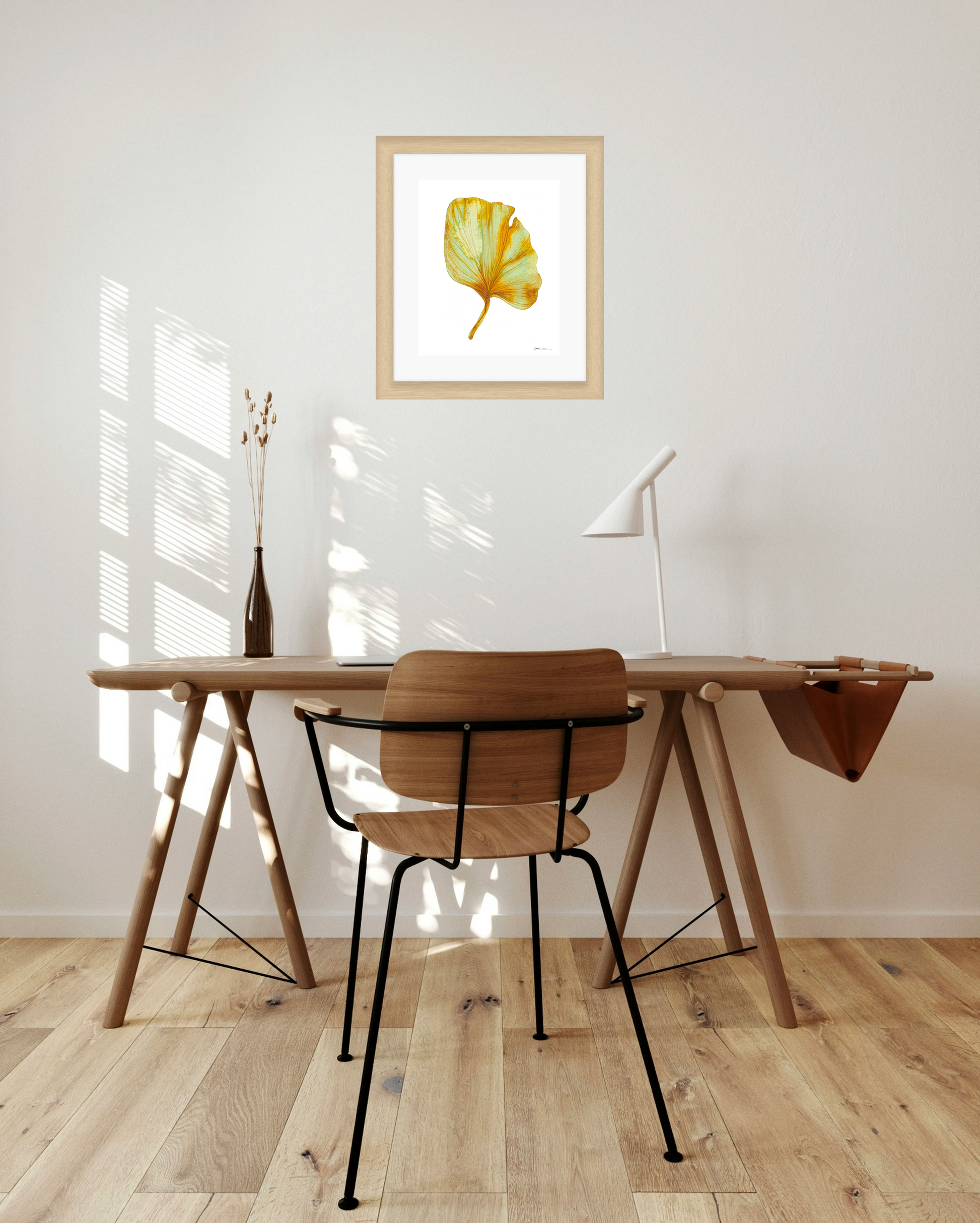 Wooden desk with chair in a room with a framed artwork of a yellow and green leaf on a white background on the wall.