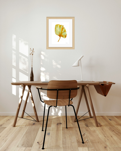 Wooden desk with chair in a room with a framed artwork of a yellow and green leaf on a white background on the wall.